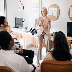Un experto en gerencia estratégica vestido formalmente da indicaciones a un equipo de trabajo desde lo alto de una mesa.